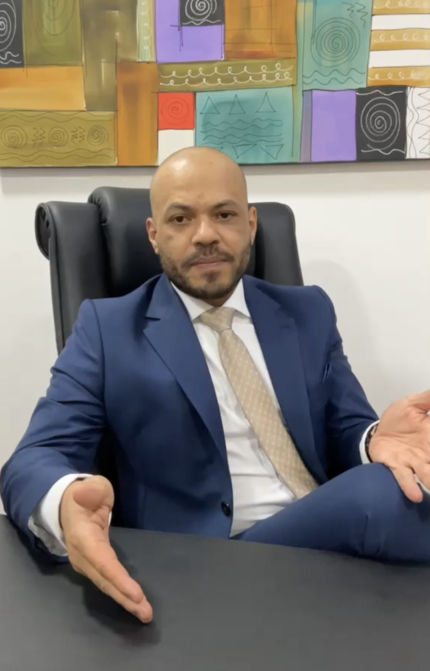 Advogado José Luiz do Carmo Chaves de terno azul marinho, sentado em cadeira de escritório executiva preta, com quadro abstrato colorido ao fundo, gesticulando com a mão em direção à mesa, retrato profissional corporativo, iluminação natural suave