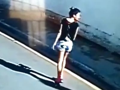 Imagem granulada de câmera de segurança mostrando uma jovem caminhando sozinha por uma via pública durante o dia, sombra projetada no chão, still usado como prova em reportagem policial