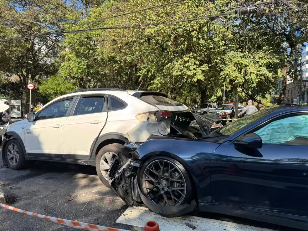 Porsche preto com a lateral dianteira destruída colidido contra a traseira de um SUV branco em via pública arborizada de bairro nobre de São Paulo, fita de isolamento laranja e branca no chão, cone de sinalização, policiais ao fundo, dia ensolarado, fotografia jornalística documental