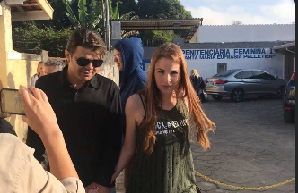 Fotografia documental de imprensa mostrando um grupo de pessoas do lado de fora de penitenciária feminina em dia ensolarado, fachada com placa 'Penitenciária Feminina Santa Maria Eufrásia Pelletier', homem de óculos escuros e mulher de cabelos longos ao centro, pessoa filmando com celular em primeiro plano, policial ao fundo, viaturas estacionadas